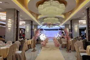 Salão de banquetes com um corredor central, mesas redondas e palco com decorações florais.