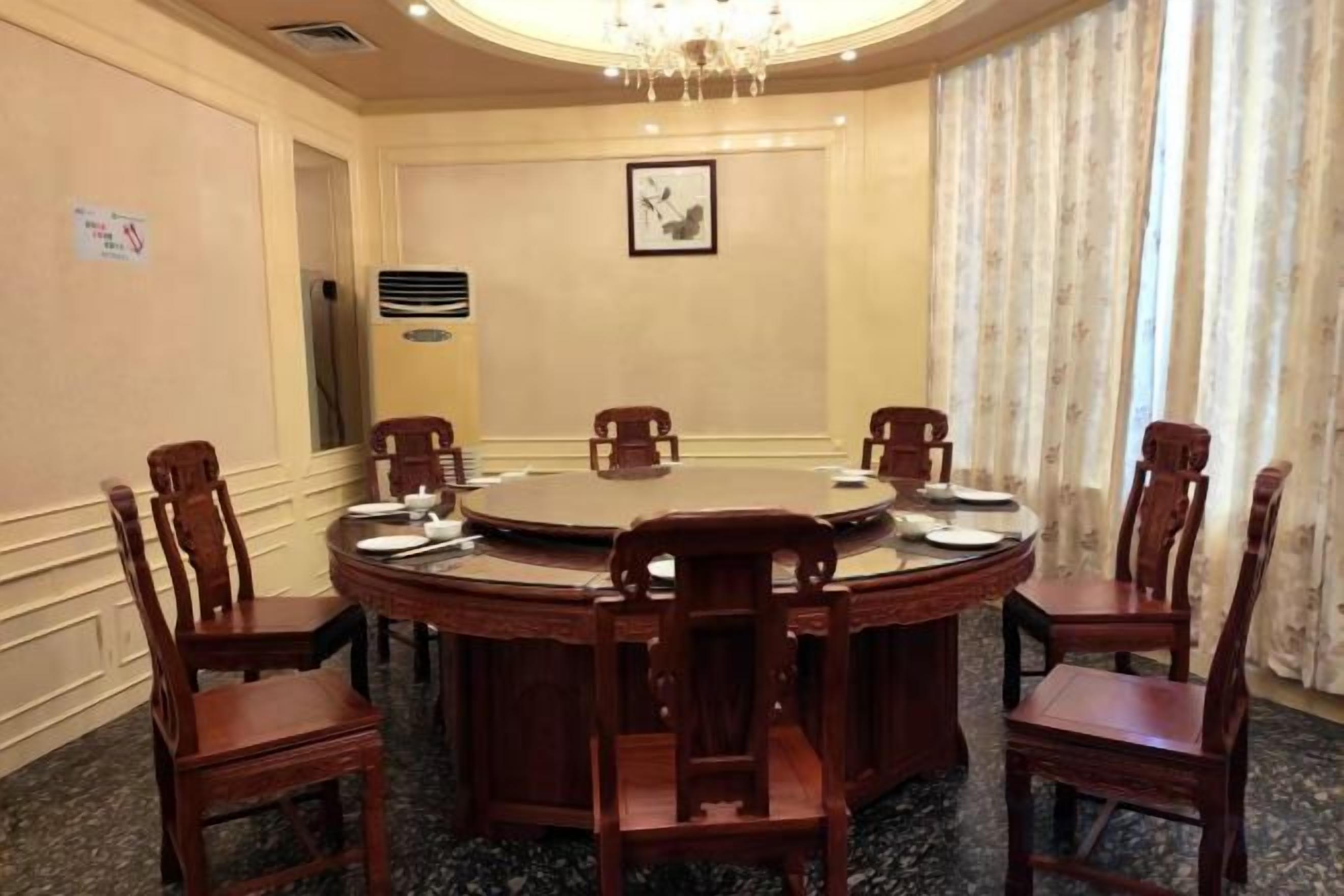 Un salón de conferencias con una mesa redonda, seis sillas de madera, una araña y un ventanal con cortinas.