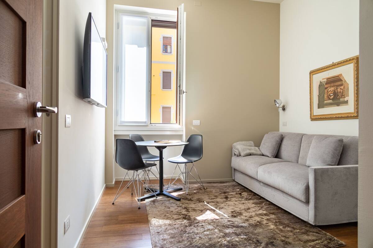 A living room with a grey sofa, a small round table with chairs, a TV mounted on the wall, and a window with a view of a building.