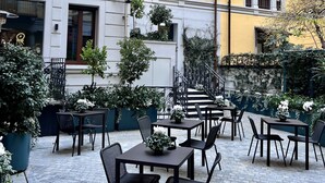 En udendørs café med borde og stole, omgivet af krukker med planter og en gul bygning med skodder.