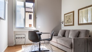 En moderne stue med en grå sofa, et spisebord med stole, et lille sofabord og et stort vindue med udsigt til bygninger.