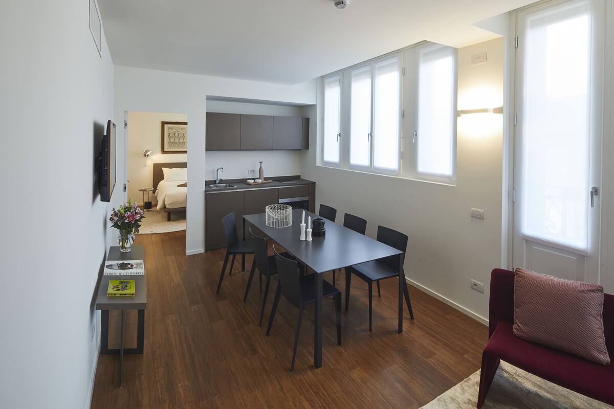 A modern kitchen with a dining area, a red sofa, and a small table with a vase of flowers.