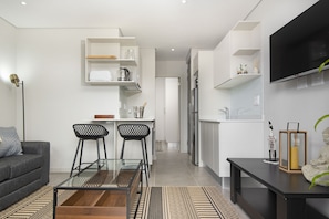A modern living space with a sofa, a glass coffee table, a kitchen area with a fridge, and a TV mounted on the wall.