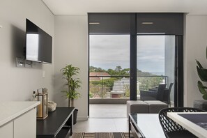 A modern living room with a flat-screen TV, a sofa, a dining table, and a balcony with a view of the city.