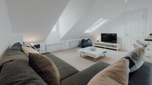A modern loft-style living room with a high ceiling, skylight, and a flat-screen TV on a white stand.