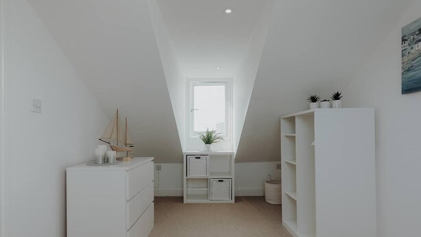 A narrow, white-walled room with a sloped ceiling, a small window, and a view of a coastal scene.