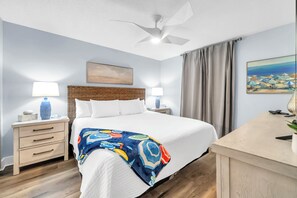 Ein Hotelzimmer mit einem großen Bett, zwei Nachttischen mit Lampen, einem Deckenventilator, einem Gemälde und Blick auf den Strand.