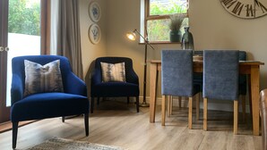 A living room with a large wall clock, a dining table with chairs, a lamp, and a window with curtains. - Come & Stay on a Real Norfolk Vineyard (Norwich)