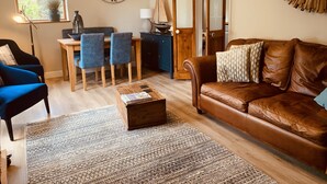 A living room with a brown leather sofa, a wooden coffee table, a blue armchair, a dining table with blue chairs, a large wall clock, and a mirror with a wooden frame. - Come & Stay on a Real Norfolk Vineyard (Norwich)