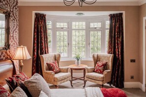 A room with two armchairs, a round table, and a sofa. There are large windows with curtains, a lamp, and a plant on the table.