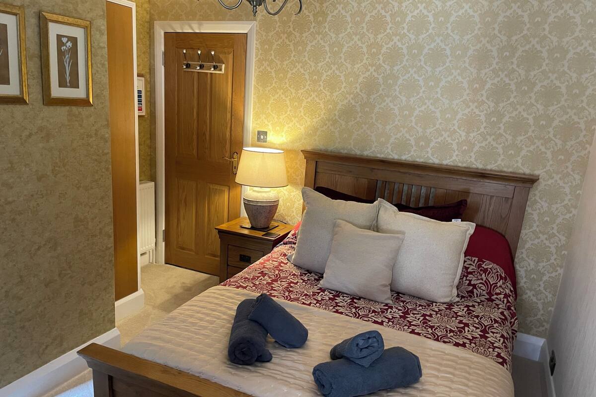 A neatly made bed with a patterned bedspread, a wooden headboard, and two rolled towels on the bed.