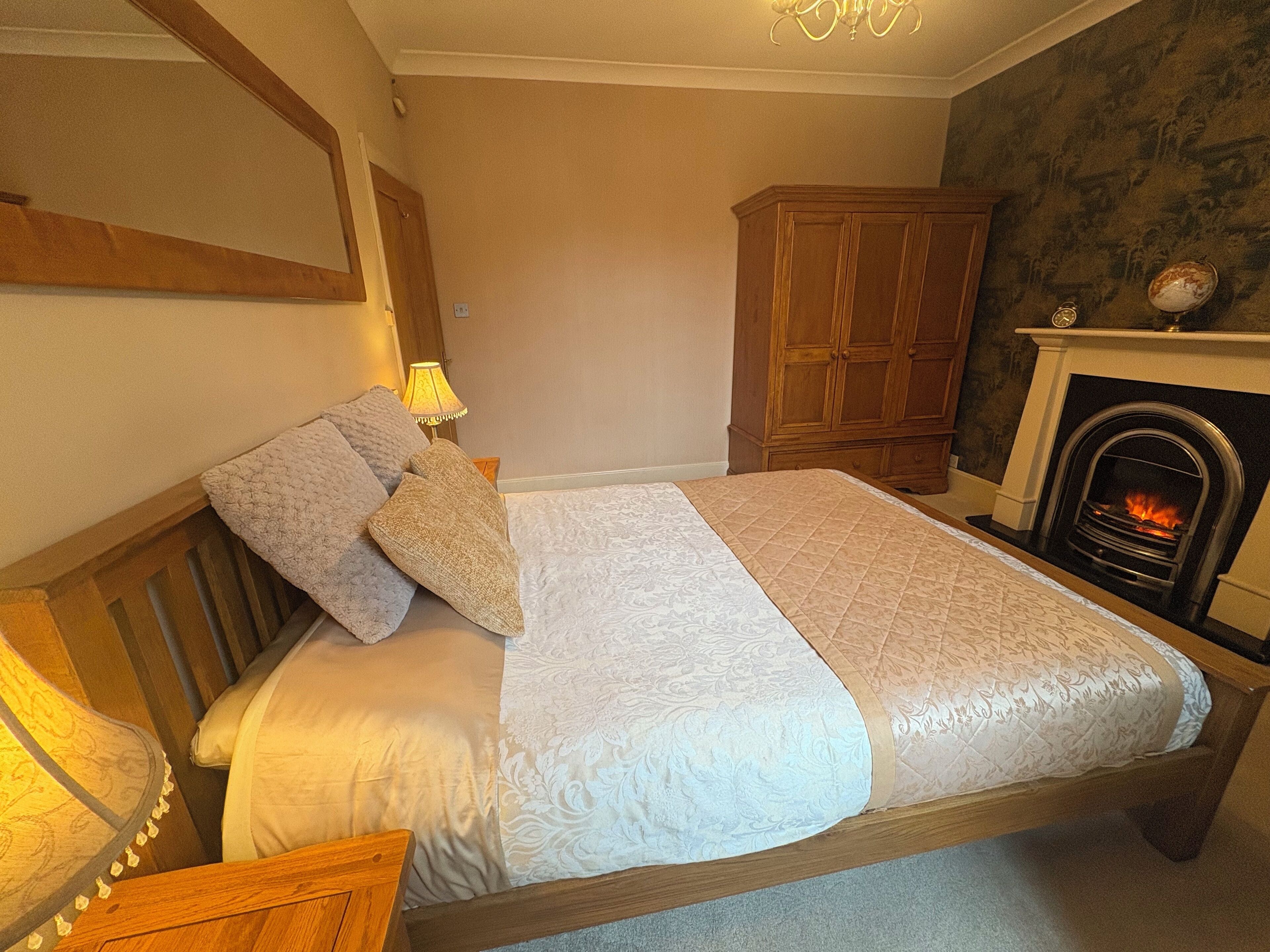 A bedroom with a bed, a wooden wardrobe, a nightstand, a lamp, and a fireplace.