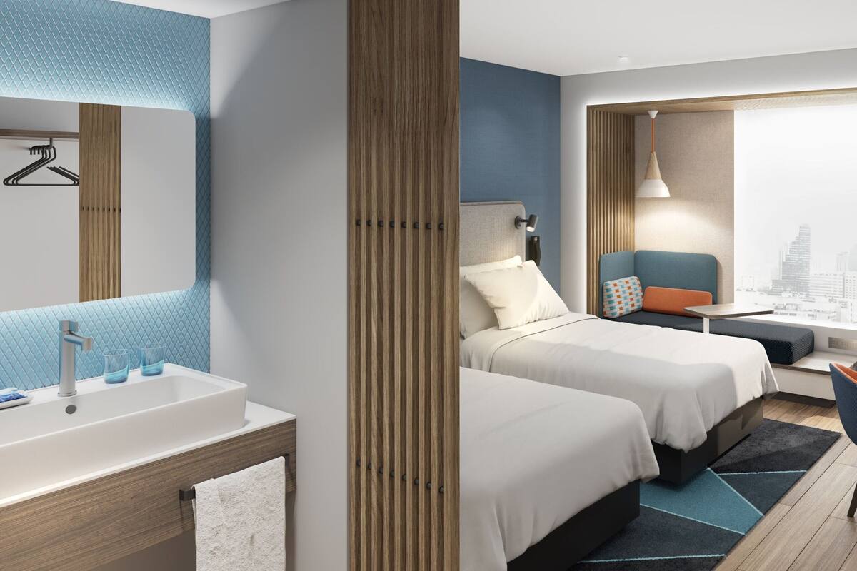 A modern hotel room with two beds, a sitting area, a desk, and a large window with a city view.