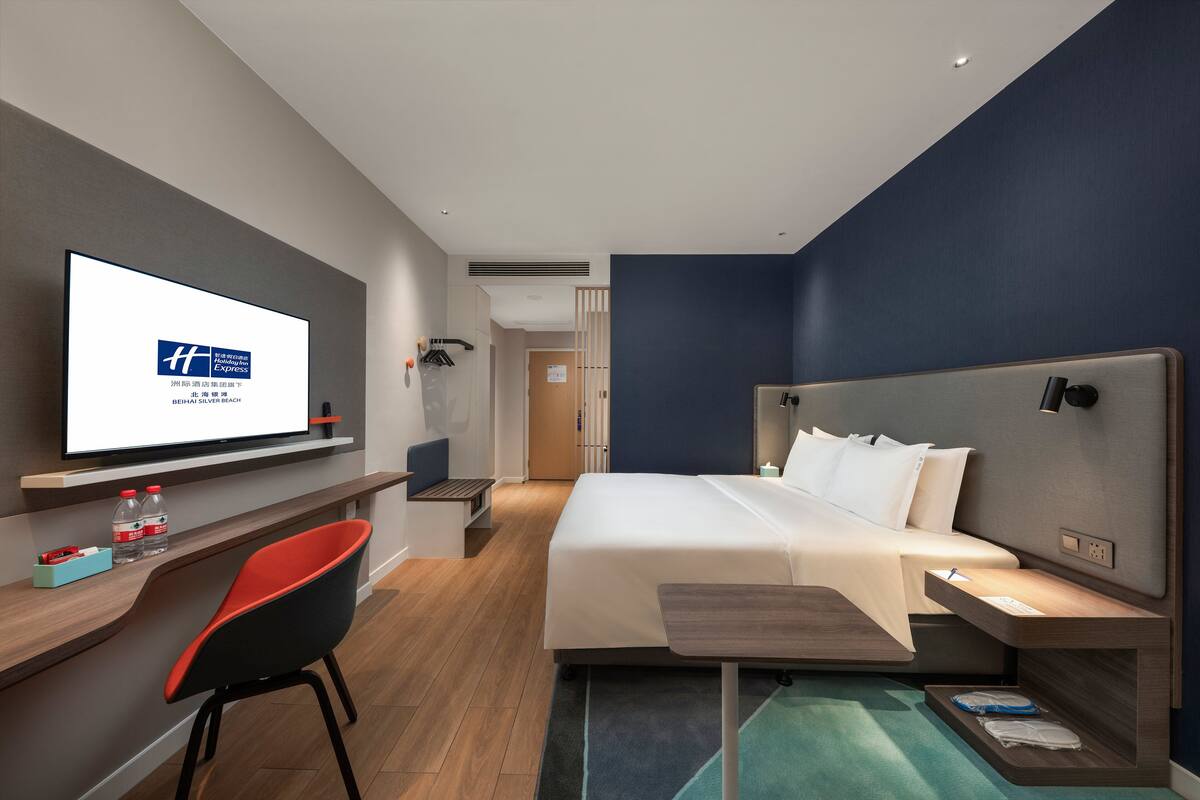 A modern hotel room with a bed, a desk with a chair, a television, and a bathroom visible in the background.