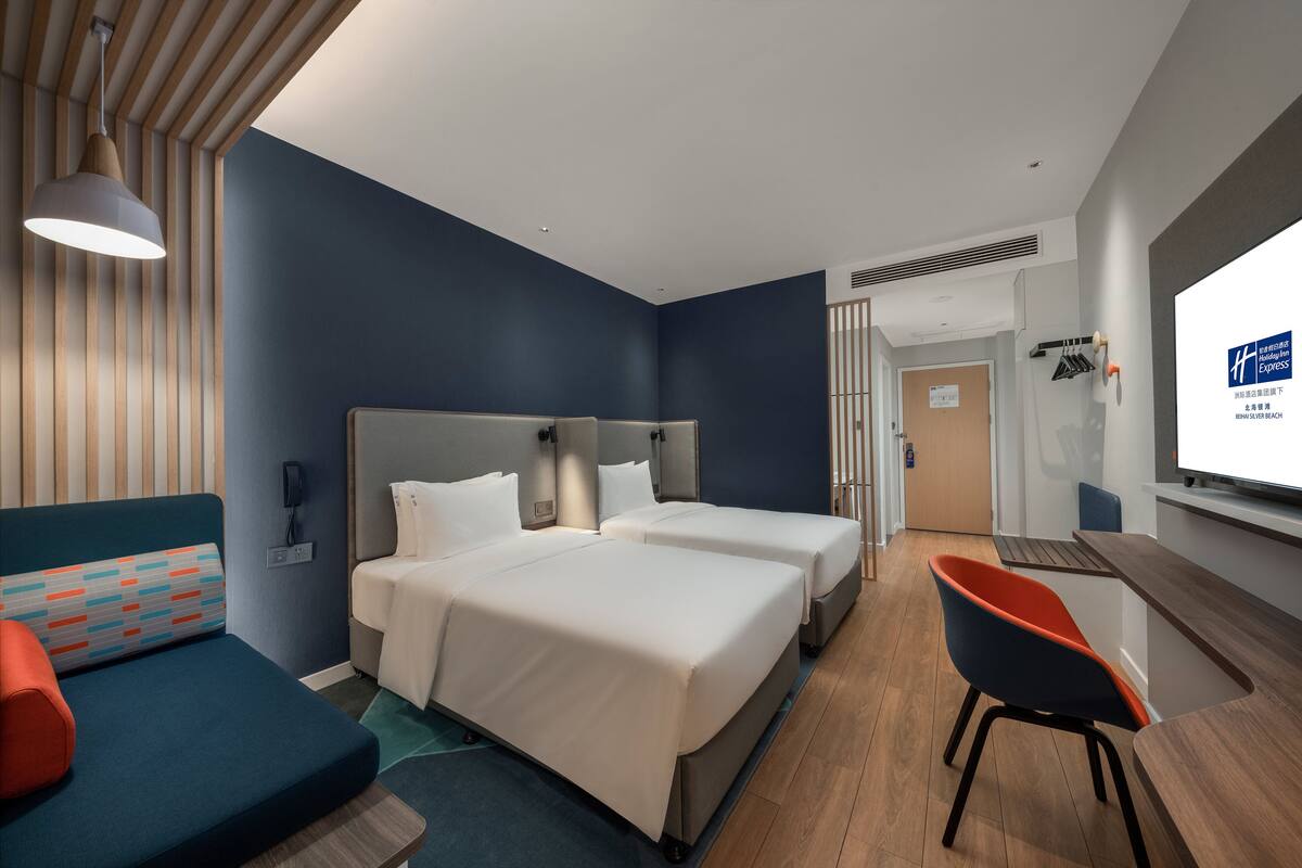 A hotel room with two beds, a desk, a chair, and a TV.