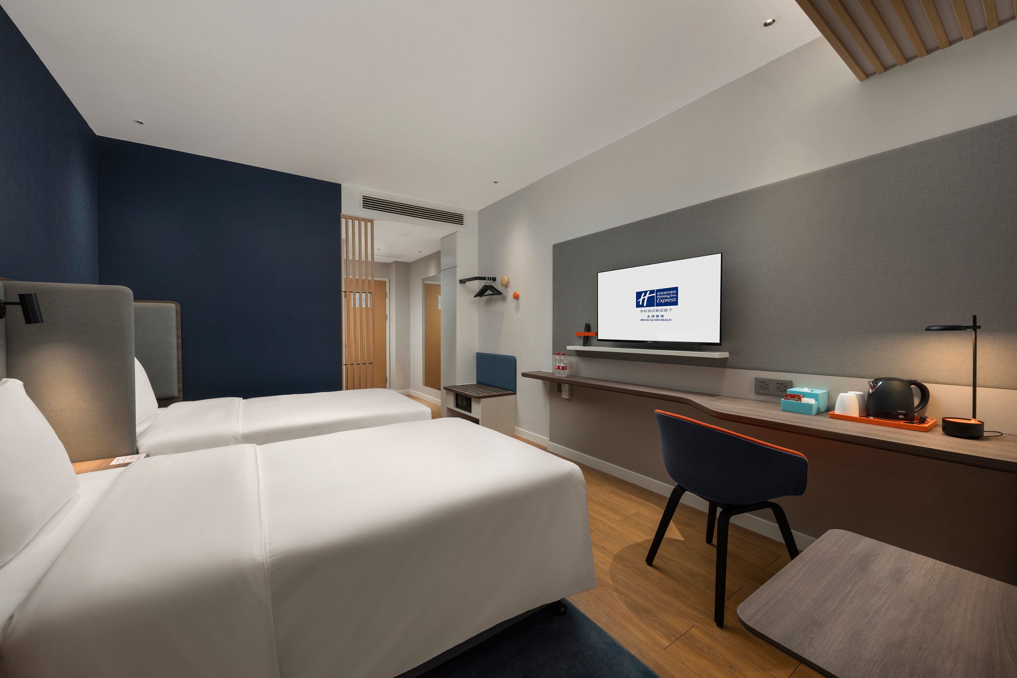 A modern hotel room with two beds, a desk, a chair, a TV, and a small kitchenette.