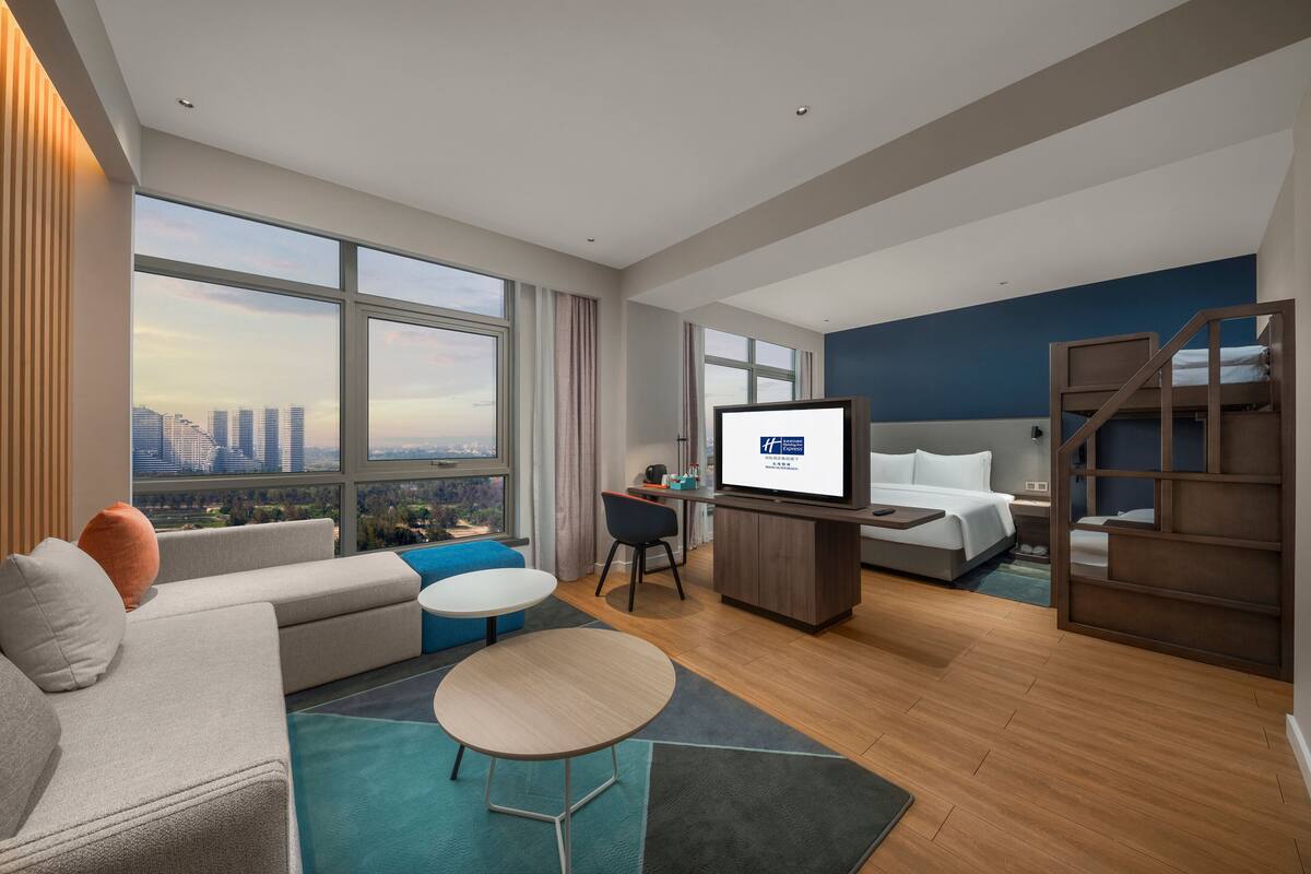A modern hotel room with a large window, a bed, a desk, and a television.