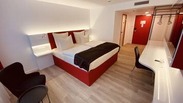 Habitación de hotel moderna con una cama de color rojo y blanco, un escritorio pequeño, dos sillas y un espejo.