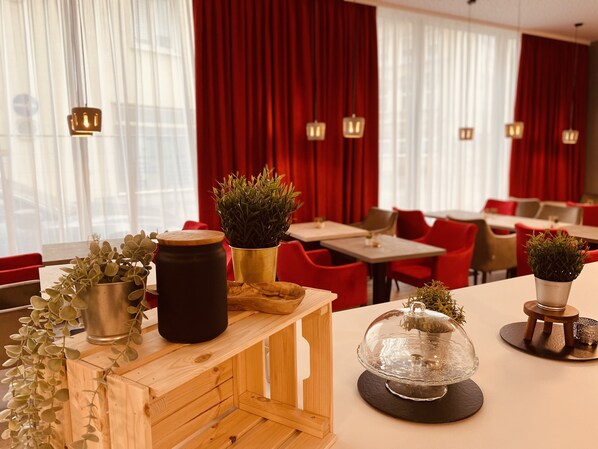 Un interior de restaurante con cortinas rojas, mesas de madera y plantas en macetas.