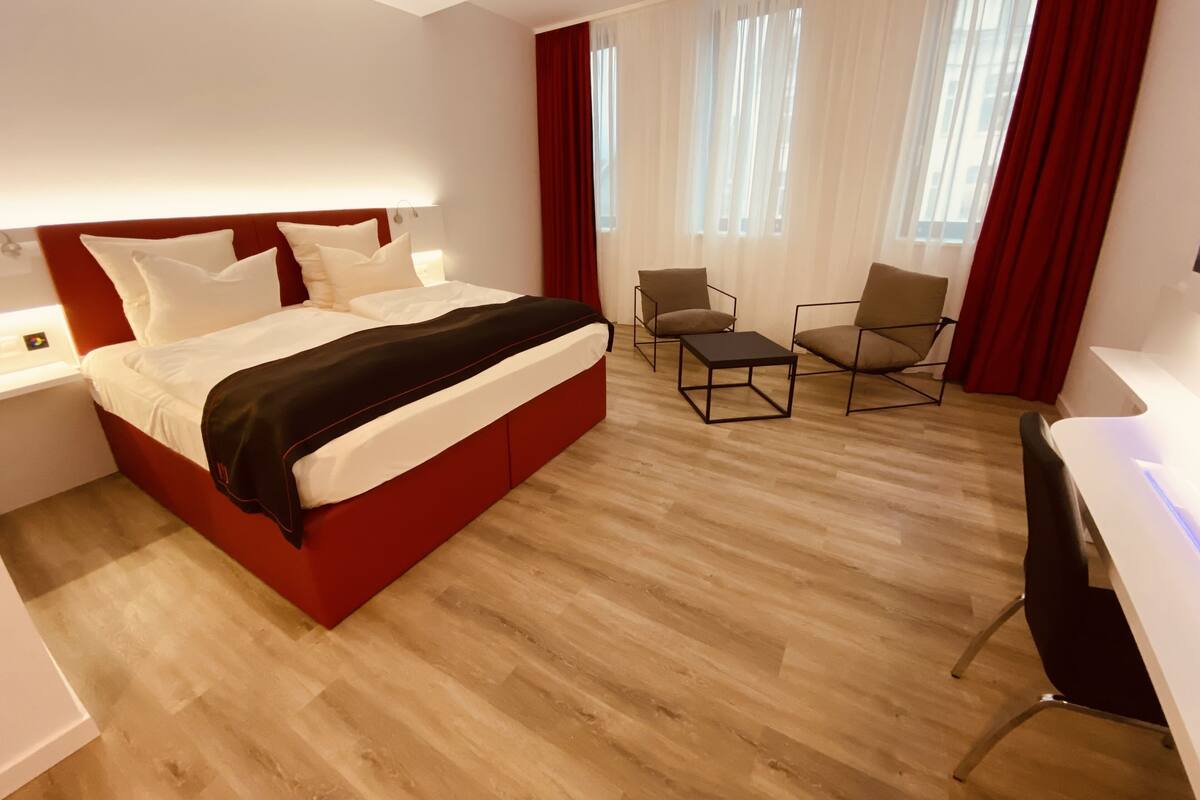 Une chambre d’hôtel moderne avec un grand lit, deux fauteuils, une petite table et un bureau.