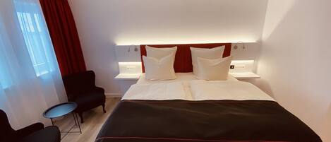 Una habitación de hotel moderna con una cama grande, una mesita de noche, una silla y una ventana con cortinas.