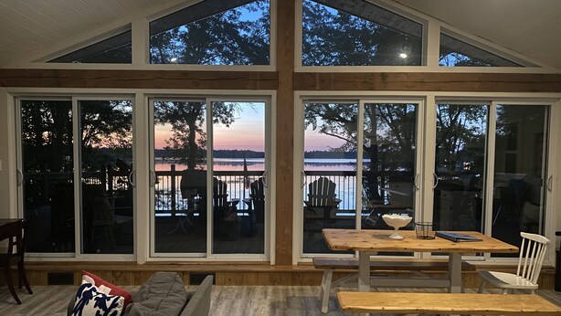 A spacious room with large windows offering views of a lake and sunset. There is a wooden table and chairs, and a sofa with cushions.