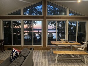 A spacious room with large windows offering views of a lake and sunset. There is a wooden table and chairs, and a sofa with cushions. - Cozy 4 Season Cottage stunning sunsets on Sparrow Lake!(Pet friendly)New Hot Tub (Severn Bridge)