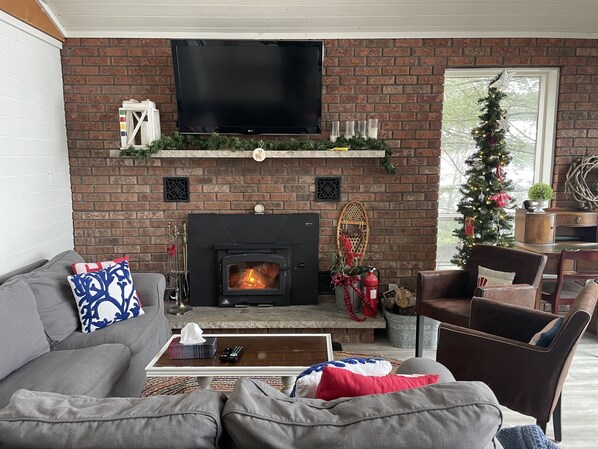 A cozy living room with a brick fireplace, a flat-screen TV, and a Christmas tree. - Cozy 4 Season Cottage stunning sunsets on Sparrow Lake!(Pet friendly)New Hot Tub (Severn Bridge)