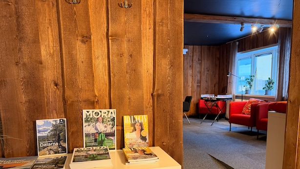 A wooden interior with a white table displaying magazines, a red sofa, and a window with plants. - Fridhemsgatan City Stay (Mora)