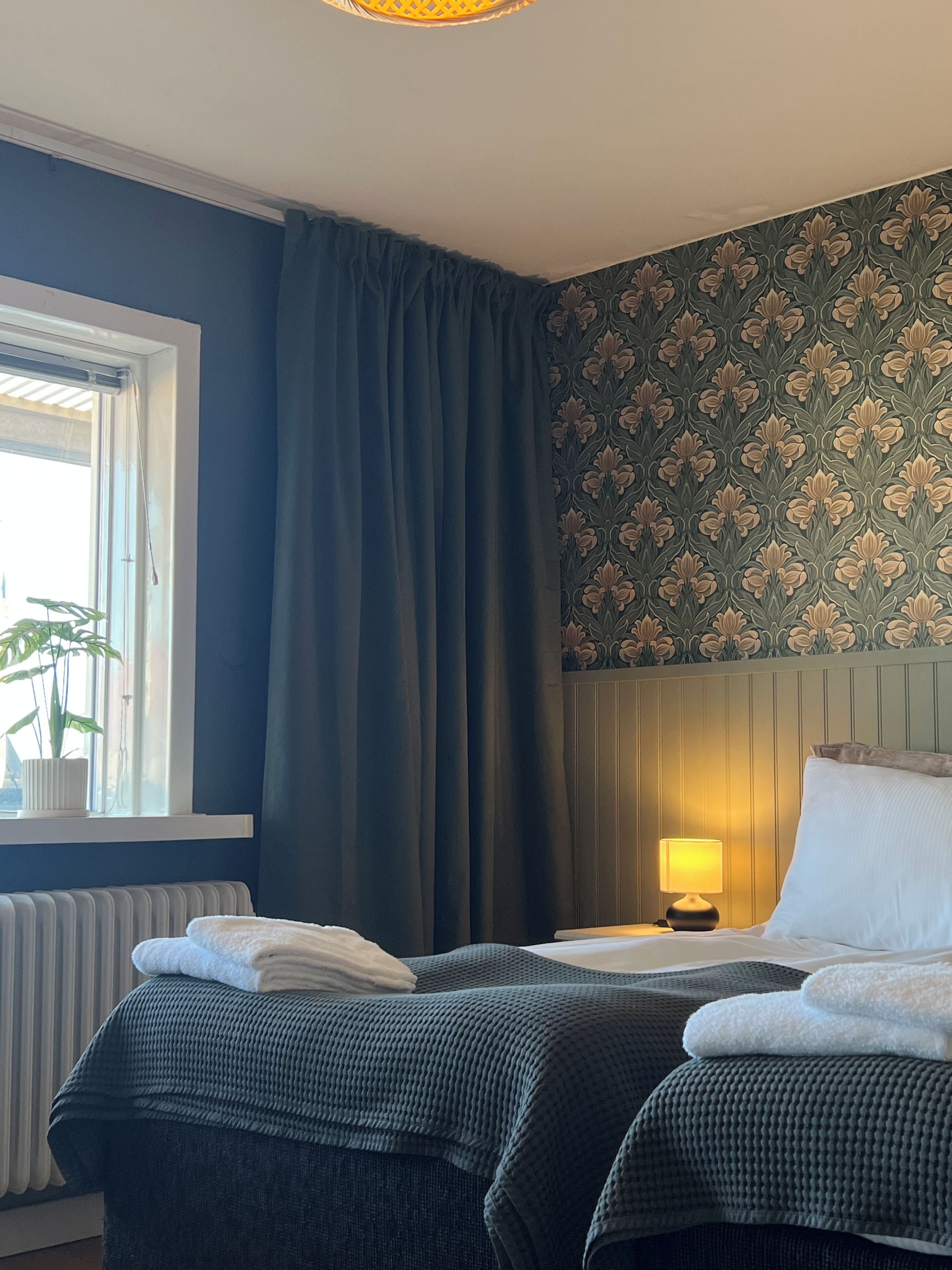 Ein Hotelzimmer mit zwei Betten, einem Fenster mit Vorhängen, einem Heizkörper, einer Wand mit Blumenmuster und einer Nachttischlampe.