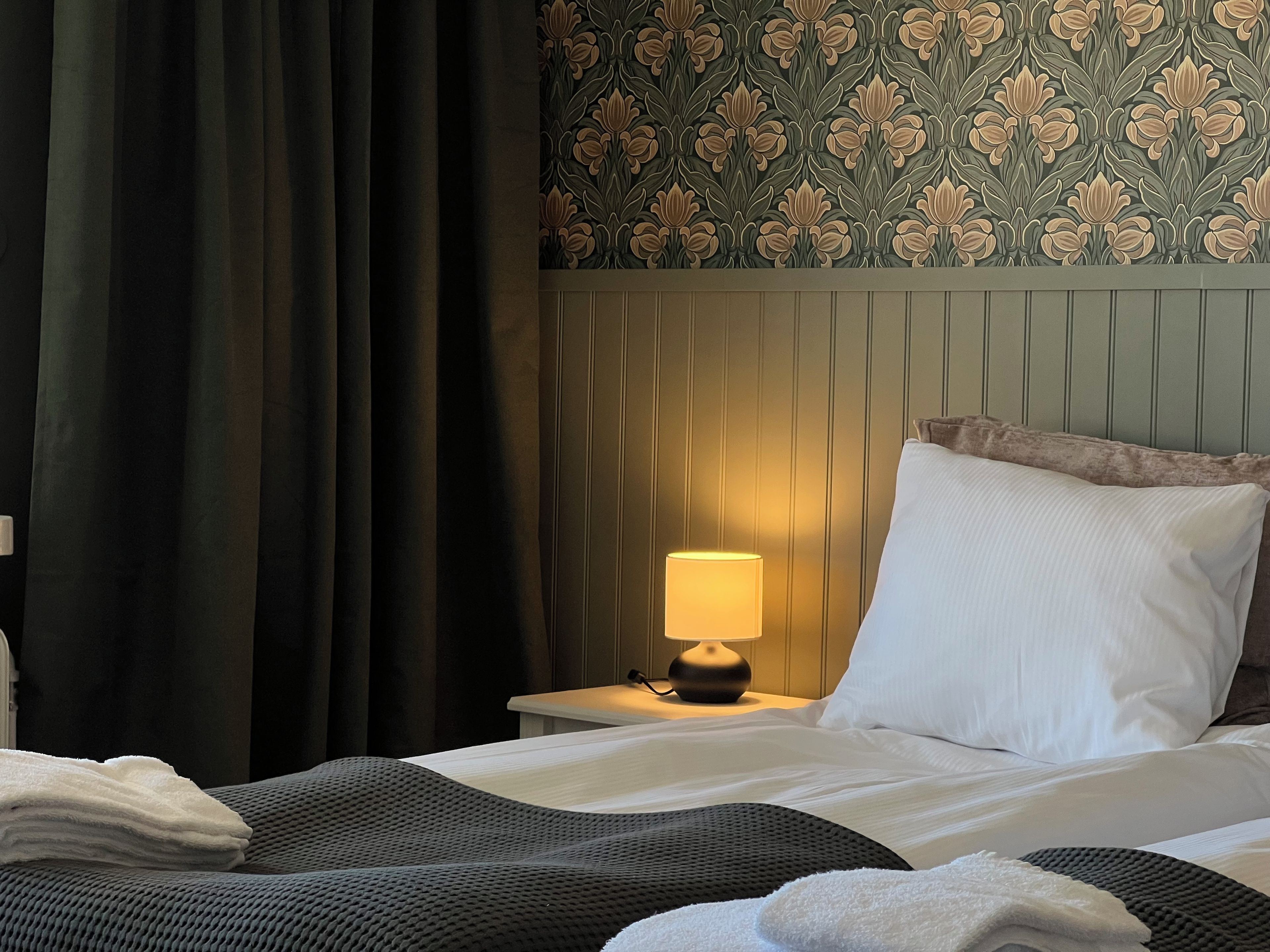A hotel room with a bed, a nightstand with a lamp, a window with curtains, and a patterned wallpaper.
