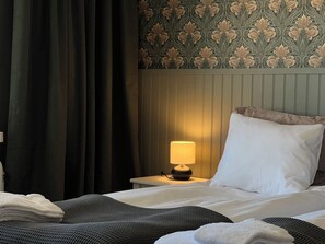 A hotel room with a bed, a nightstand with a lamp, a window with curtains, and a patterned wallpaper.