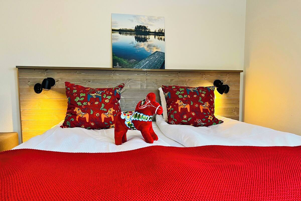 Una cama con cabecera de madera, dos almohadones decorativos, una colcha roja y un cuadro de un lago.