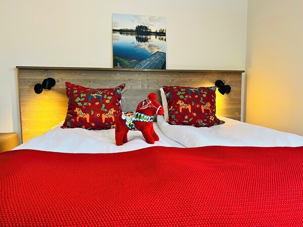 Uma cama com cabeceira de madeira, duas almofadas decorativas, um cobertor vermelho e um quadro de um lago.