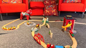 A toy train set on a carpet with a wooden floor, surrounded by red sofas with floral patterns. - Fridhemsgatan City Stay (Mora)