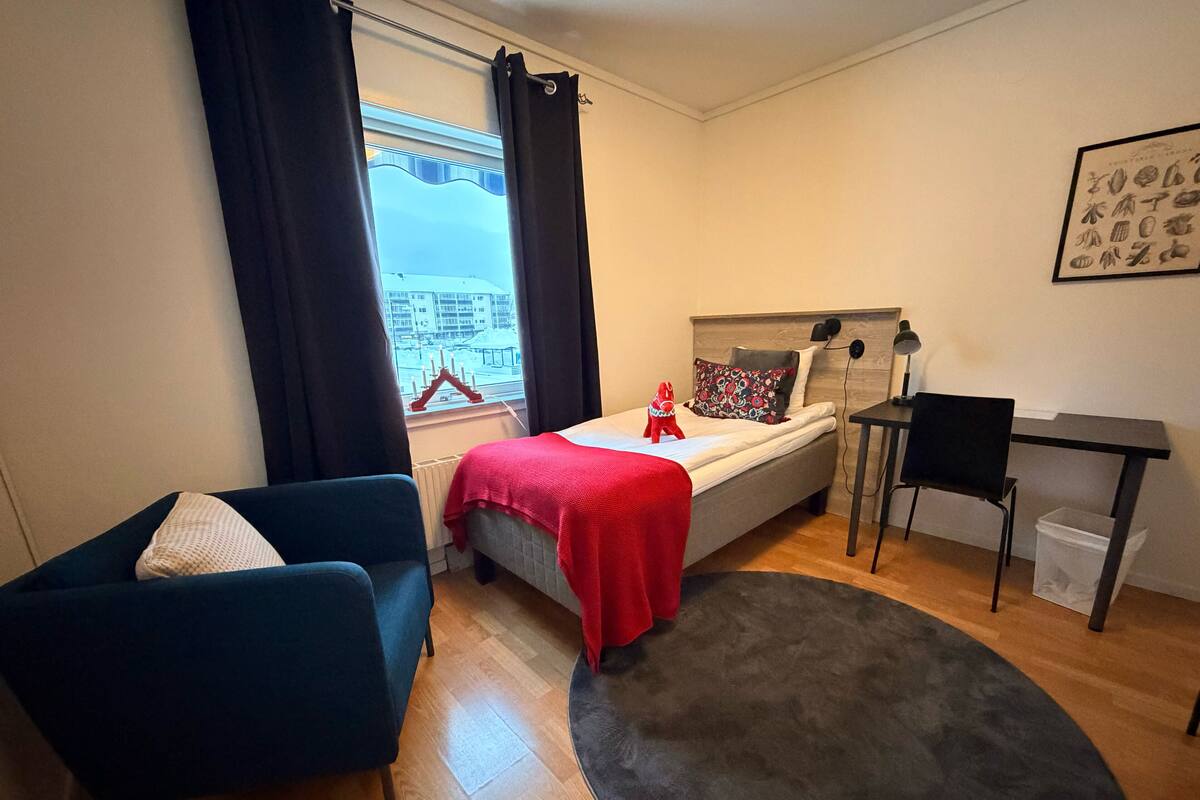 Un dormitorio compacto con cama, escritorio, silla, sillón azul, ventana con cortinas y vista a edificios.