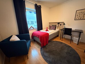 Quarto compacto com cama, escrivaninha, cadeira, poltrona azul, janela com cortinas e vista para edifícios.