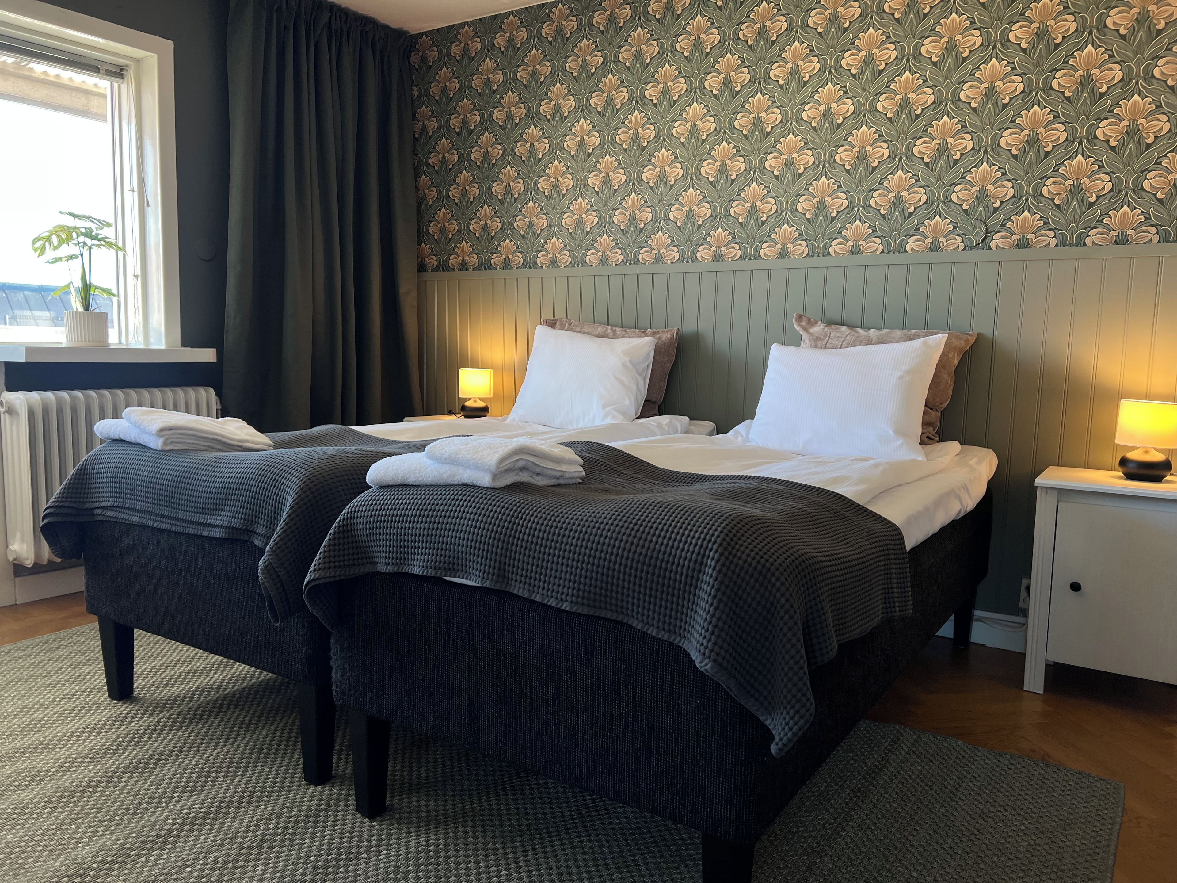 A hotel room with a double bed, two bedside tables with lamps, a radiator, a window with curtains, and a floral wallpaper pattern.