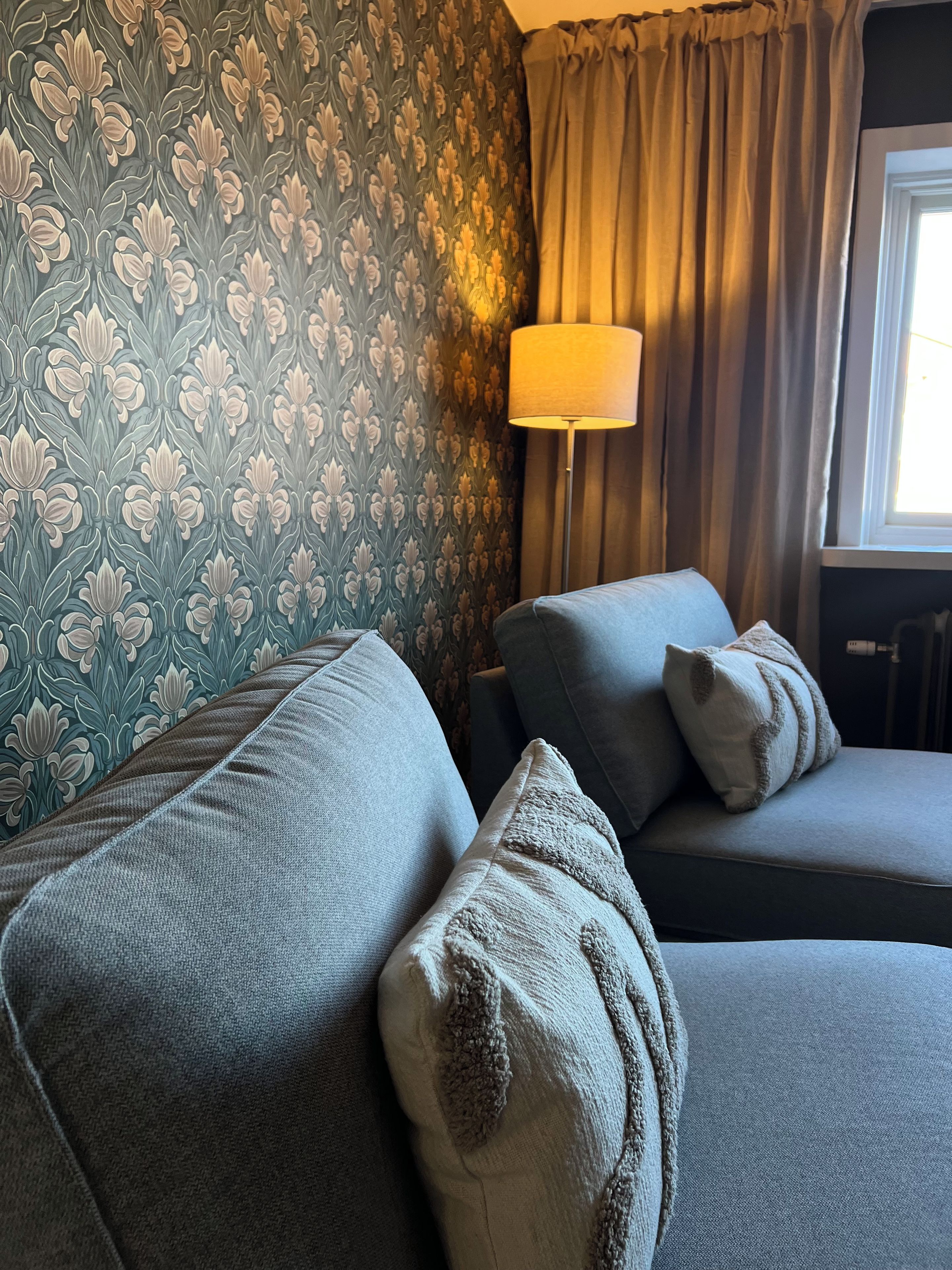 Una habitación con papel tapiz estampado, un sofá con almohadones, una lámpara de pie y un ventanal con cortinas.