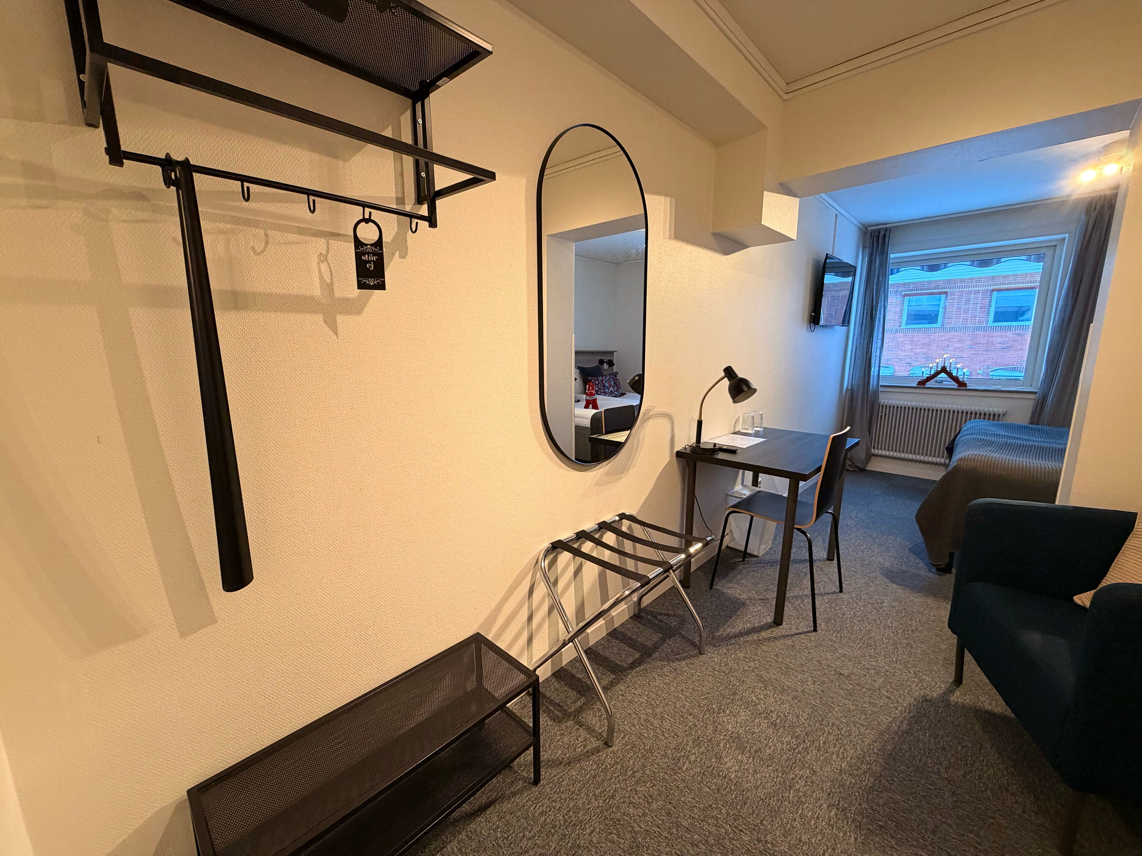 Habitación de hotel compacta con cama, escritorio con silla, mesita pequeña, espejo y perchero.