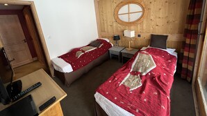A room with two single beds, a wooden wall, a round window, a small table with a TV and remote controls, and a door.