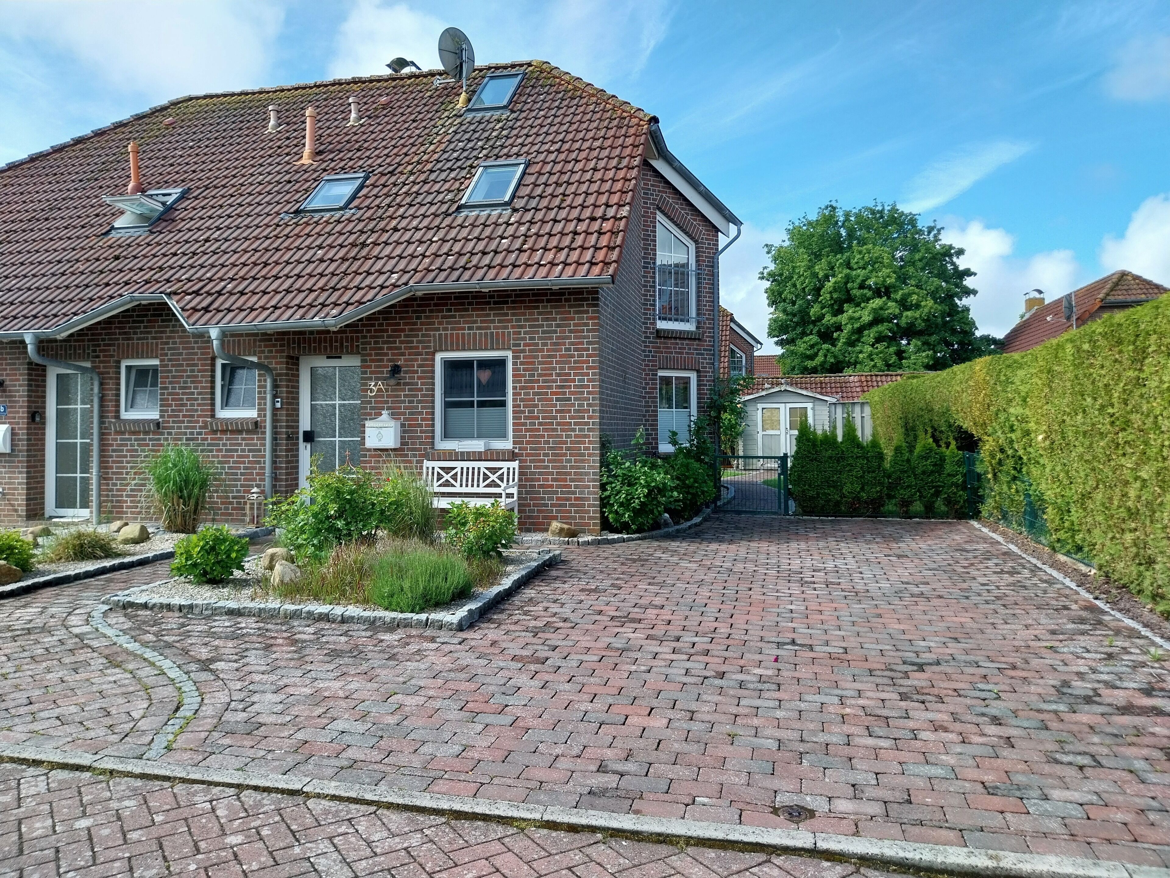 Een huis van baksteen met twee verdiepingen, voorzien van een pannendak, een schotelantenne en een kleine tuin met een bankje.