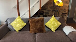Uma sala de estar com um sofá marrom, duas almofadas amarelas com bolinhas e um travesseiro fofo marrom. Uma parede de pedra e uma escada de madeira são visíveis ao fundo.