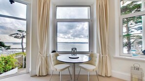 Een kamer met een groot raam, twee stoelen, een ronde tafel en uitzicht op zee.