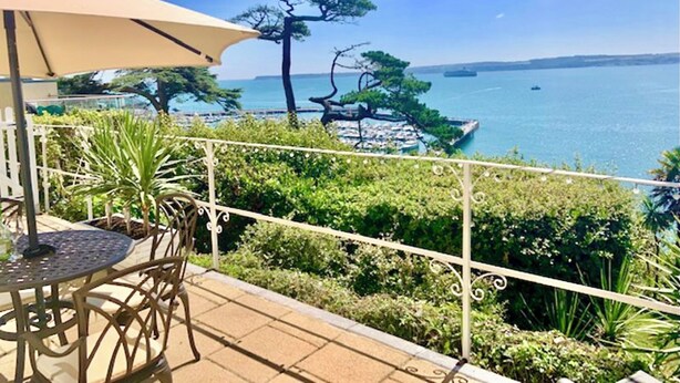 Een balkon met een tafel en stoelen, uitkijkend op een kustgebied met bomen en uitzicht op zee.