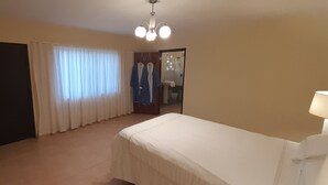 Un dormitorio con cama, puerta, ventana con cortinas, espejo y lámpara en el techo.