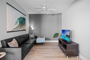 A modern living room with a black leather sofa, a wooden coffee table, a TV on a dark wooden stand, and a ceiling fan.