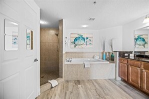 A modern bathroom with a bathtub, a shower, and a walk-in shower. There are framed artworks on the walls and a wooden floor. - #702 Sanibel - Oceanfront Condo Panoramic Views (Daytona Beach Shores)