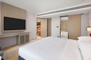 A hotel room with a large bed, a flat-screen TV mounted on the wall, a seating area with chairs, and a bathroom visible through an open door.