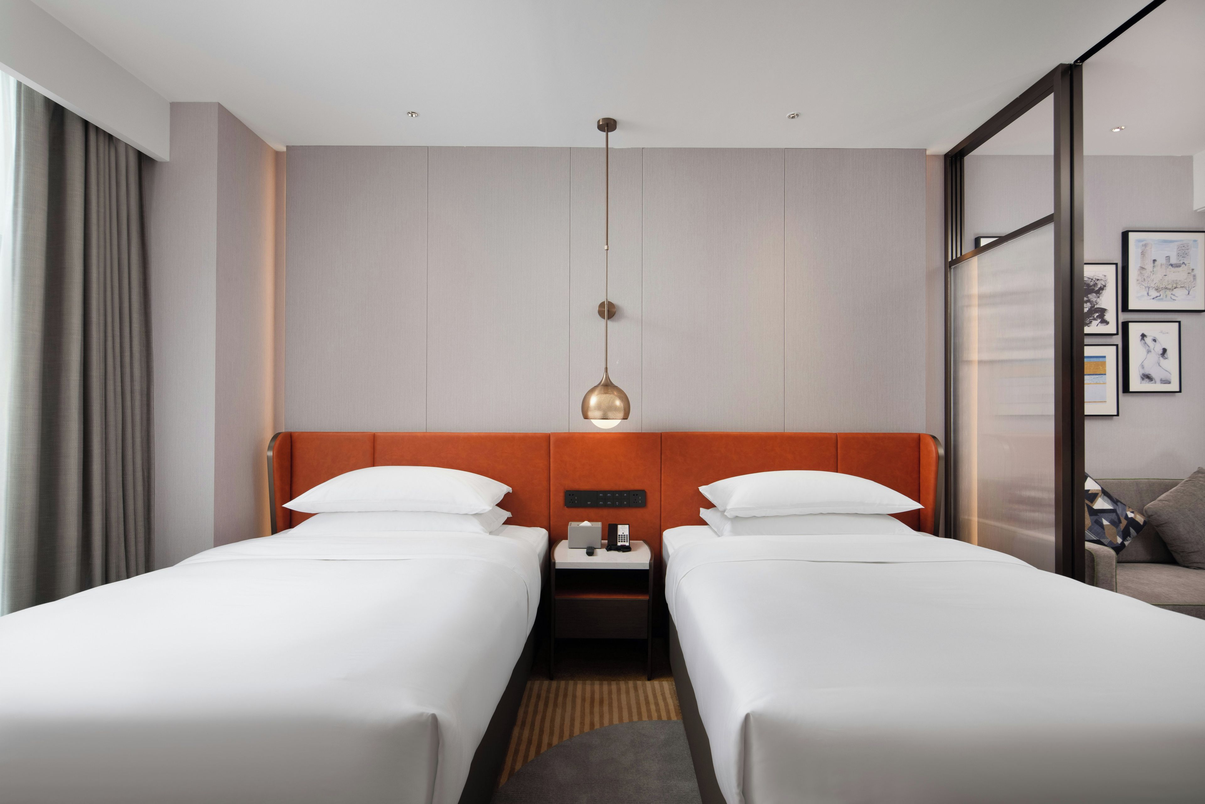 Quarto de hotel com duas camas, uma mesa de cabeceira, uma luminária e uma janela com cortinas.